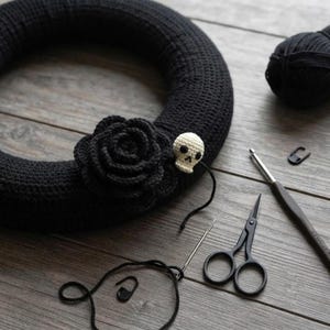 May include: A black crocheted wreath base adorned with a large black crocheted rose and a small cream-colored crocheted skull. The scene includes a ball of black yarn, a metal crochet hook, black scissors, a sewing needle, and two stitch markers.