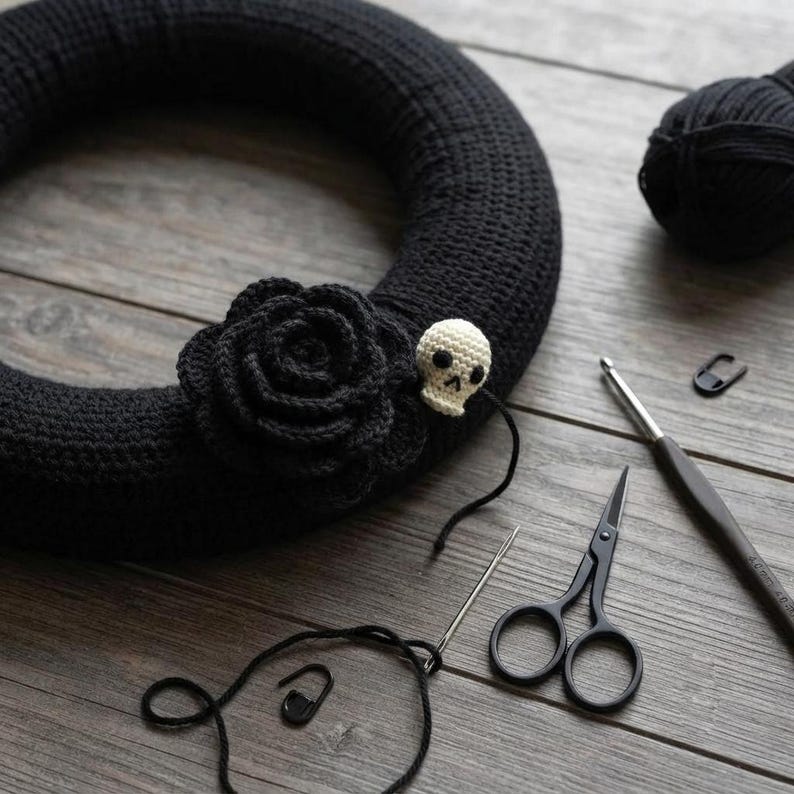 May include: A black crocheted wreath base adorned with a large black crocheted rose and a small cream-colored crocheted skull. The scene includes a ball of black yarn, a metal crochet hook, black scissors, a sewing needle, and two stitch markers.