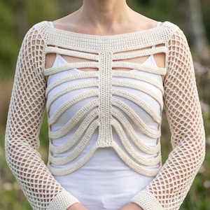 May include: A cream-colored crochet bolero featuring a ribcage-inspired design with horizontal bands across the chest and long sleeves with an open net pattern. The garment is worn over a plain white tank top, showcasing the intricate stitch work.