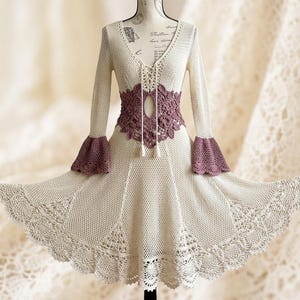 Può includere: Un abito all'uncinetto color crema con maniche lunghe e polsini svasati, scollo a V profondo con allacciatura e un pannello floreale all'uncinetto color malva in vita. La gonna ha una silhouette svasata con motivi in pizzo sull'orlo.