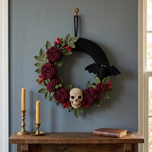 Puede incluir: Una corona circular con una base de ganchillo negra adornada con rosas de ganchillo de color rojo oscuro, hojas verdes, pequeñas bayas rojas, una calavera de ganchillo beige en el centro inferior y un murciélago de ganchillo negro a la derecha.