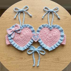 May include: A handmade crochet top featuring two large pink heart-shaped cups with light blue ruffled edges. The garment includes light blue tie-up shoulder straps, a small central heart detail, a pink bow on the left cup, and a light blue tie closure.