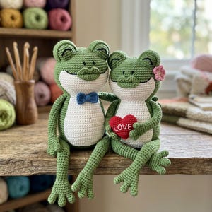 Peut inclure: Deux grenouilles crochetées vertes avec des ventres blancs assises côte à côte. La grenouille de gauche porte un petit nœud papillon bleu. La grenouille de droite tient un coussin rouge en forme de cœur avec le mot LOVE brodé en blanc.