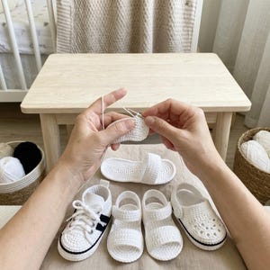 May include: A pair of hands crocheting a white shoe with a silver hook. Below are four finished white crochet shoes: a sneaker with black accents, two sandals with single straps, and a clog-style shoe with a black trim along the sole.