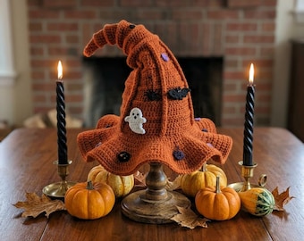 May include: A crocheted orange witch hat featuring small appliqués of a white ghost, black bats, and purple circles. The hat is displayed on a wooden stand surrounded by several small orange pumpkins and two black taper candles in brass holders.