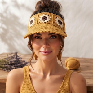 May include: A mustard yellow crochet visor featuring three prominent sunflower motifs across the front. The sunflowers have white petals and dark brown centers, crafted with a textured yarn stitch. The visor has a wide, structured brim.