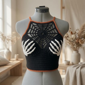 May include: A black crochet crop top featuring a white skeleton hand design on the chest and a black spiderweb pattern across the upper neckline. The garment is finished with thin orange trim along the neckline and the bottom hemline.