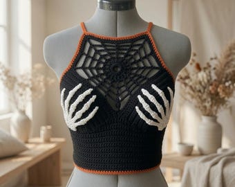 May include: A black crochet crop top featuring a white skeleton hand design on the chest and a black spiderweb pattern across the upper neckline. The garment is finished with thin orange trim along the neckline and the bottom hemline.