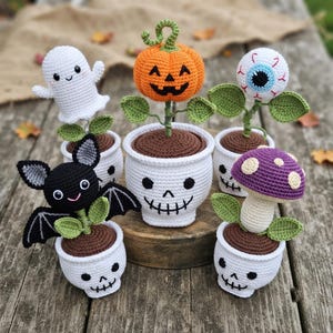May include: A collection of five crocheted items in white skull-shaped pots. The set includes a white ghost, an orange jack-o'-lantern, a blue and white eyeball, a black bat with grey ears, and a purple mushroom with white spots on its cap.