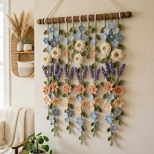 May include: A wall hanging featuring crocheted flowers and vines suspended from a dark wooden rod. The arrangement includes blue, cream, peach, and purple blossoms with green leaves, all crafted from yarn and hanging in vertical strands.
