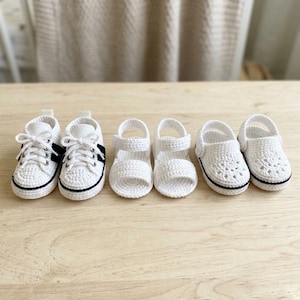 May include: A set of three pairs of handmade crochet baby shoes. The first pair are white sneakers with black accents and laces. The second pair are white open-toe sandals with a strap. The third pair are white clogs with a perforated design.