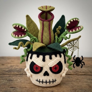 May include: A crocheted plant arrangement featuring a central pitcher plant and two Venus flytraps with green and maroon leaves. The plants are housed in a cream-colored skull-shaped pot with red button eyes and a hanging black spider web.
