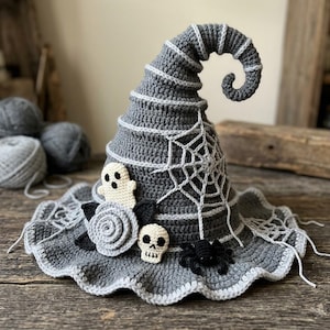 Puede incluir: Un sombrero de bruja de ganchillo gris con punta curvada, decorado con un patrón de telaraña blanca, un pequeño fantasma blanco, una rosa gris claro, una hoja negra, una pequeña calavera blanca y una araña negra en el ala ancha y ondulada.