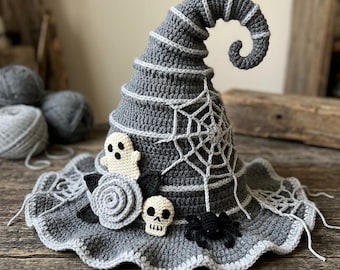 May include: A gray crocheted witch hat featuring a curled tip, decorated with a white spiderweb pattern, a small white ghost, a light gray rose, a black leaf, a small white skull, and a black spider attached to the wide, wavy brim.