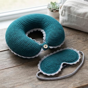 Puede incluir: Una almohada de viaje de ganchillo en color verde azulado oscuro con un borde festoneado gris claro y un cierre de botón de madera, junto con un antifaz de ganchillo a juego con borde festoneado y una correa de ganchillo en gris claro.