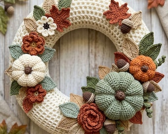 May include: A circular wreath made of cream-colored crocheted mesh, decorated with crocheted pumpkins in white, orange, and sage green, along with crocheted flowers, brown acorns, and various leaves in shades of green, tan, and rust orange.