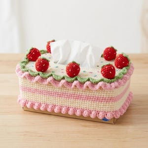 May include: A rectangular crochet tissue box cover featuring a cream and pink striped base with a scalloped pink bottom edge. The top is decorated with small red crocheted strawberries, a green ruffled border, and colorful embroidered dots.