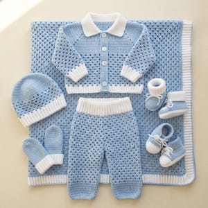 May include: A light blue and white crochet baby set featuring a long-sleeve button-up cardigan, pants, a beanie, a pair of mittens, and two pairs of booties, all arranged on a matching light blue and white crochet blanket with a border.