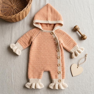 May include: A peach-colored crochet hooded romper featuring a side-button closure with eight wooden buttons. The garment includes cream-colored ribbed cuffs on the sleeves and legs, finished with delicate ruffled edges at the wrists and ankles.
