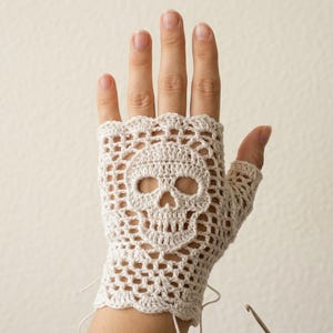 May include: A fingerless glove made of off-white crochet yarn featuring an intricate openwork pattern. The center of the glove displays a prominent skull motif integrated into the lace design, with a scalloped edge at the wrist and finger openings.