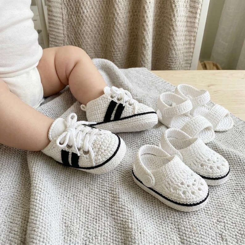 May include: A pair of white crocheted baby sneakers with black side stripes and white laces, accompanied by two additional pairs of white crocheted baby sandals featuring open-toe designs and black trim details on a textured gray fabric surface.