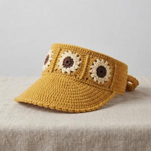 May include: A mustard yellow crochet visor featuring three prominent sunflower motifs across the front band. The sunflowers are crafted with dark brown centers and cream-colored petals, set against the textured, solid yellow yarn of the visor.