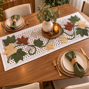 Op de afbeelding: Een witte gehaakte tafelloper met een decoratief rankpatroon en gestructureerde esdoornbladeren in de kleuren donkergroen, roestoranje en crème. De loper ligt op een houten tafel met twee couverts, elk bestaande uit een bord en een servet.