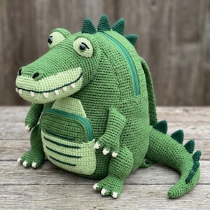 Può includere: Uno zaino a forma di coccodrillo realizzato in filato lavorato all'uncinetto verde. La borsa presenta una grande testa con denti bianchi, occhi verde scuro, uno scomparto con cerniera sul davanti e una lunga coda con punte lungo la schiena.