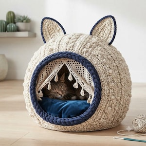Pode incluir: Uma cama redonda para animais de estimação, bege, em malha trançada, com formato de cabeça de gato e duas orelhas pontudas. A entrada é contornada com fio azul escuro e possui uma cortina de renda creme com borlas. Um gato repousa no interior.