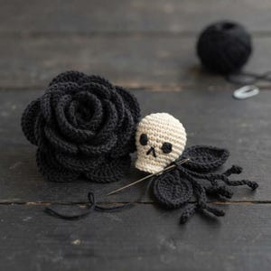 May include: A crocheted black rose flower positioned next to a small cream-colored crocheted skull with black embroidered eyes and nose. A silver sewing needle is inserted into the base of the skull, with black yarn and leaves attached.