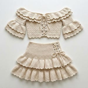 May include: A cream-colored two-piece crochet set consisting of an off-the-shoulder crop top and a matching skirt. The top features ruffled sleeves, a lace-up front with metal eyelets, and scalloped edges. The skirt has a side lace-up detail.