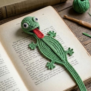 Op de afbeelding: Een groene gehaakte hagedis boekenlegger met een grote kop met witte en zwarte ogen, een lange rode tong die uit de open mond steekt, en een plat lichaam met vier kleine pootjes dat uitloopt in een lange staart op een open boekpagina.
