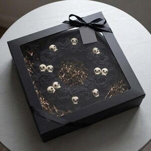May include: A square black gift box with a clear window containing a circular wreath made of black crocheted roses and small cream-colored skulls. The box is tied with a black ribbon and features a blank black gift tag attached to the top.