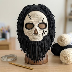 Op de afbeelding: Een gehaakt schedelmasker met een gebroken wit gezicht, zwarte gestikte scheurdetails en zwart garen in de vorm van haar en een baard. Het masker staat op een houten standaard, met een meetlint en een haaknaald op de ondergrond.