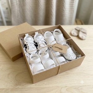 May include: A set of white crocheted baby footwear including sneakers and sandals arranged inside a square brown cardboard gift box. The items are tied with thin twine and include a small brown paper tag. Two additional white booties sit nearby.
