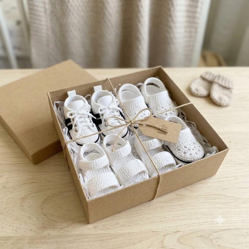 May include: A set of white crocheted baby footwear including sneakers and sandals arranged inside a square brown cardboard gift box. The items are tied with thin twine and include a small brown paper tag. Two additional white booties sit nearby.