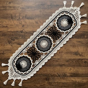 May include: An elongated oval crochet table runner featuring a cream-colored border with tassels. The center displays alternating circular web motifs in dark gray and black, connected by black spiderweb patterns, creating a detailed lace design.