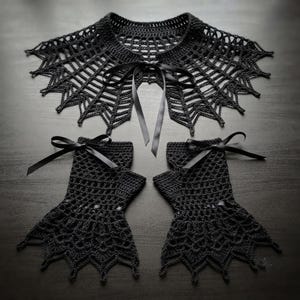 May include: A black crocheted collar and a pair of matching fingerless gloves. The collar features a web-like pattern with a central black ribbon tie. Each glove has a matching black ribbon bow at the wrist and a flared, pointed edge design.