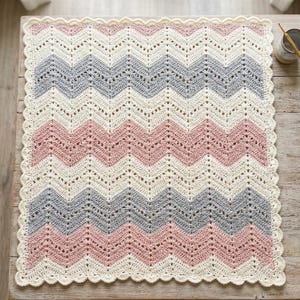 Peut inclure: Une couverture rectangulaire en crochet présentant un motif en chevron avec des rayures alternées de couleur crème, vieux rose et gris clair. La couverture possède une bordure festonnée crème sur les quatre côtés, avec un point ajouré.