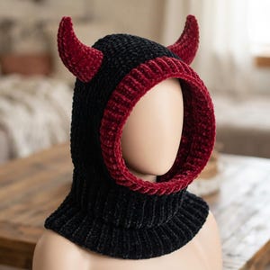 May include: A black velvet balaclava featuring a deep red ribbed trim around the face opening and two matching red velvet horns protruding from the top. The head covering has a thick, textured knit construction that extends down to the neck area.