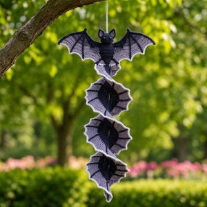 Puede incluir: Una decoración colgante de ganchillo que presenta un murciélago gris oscuro en la parte superior con las alas extendidas, conectado a una serie vertical en espiral de tres segmentos de alas adicionales en tonos alternos de gris oscuro y claro.