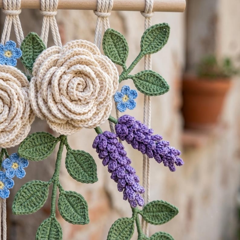 May include: A macrame wall hanging featuring crocheted cream-colored roses, small blue forget-me-not flowers, and purple lavender sprigs. The floral arrangement is accented with green crocheted leaves and suspended from a light wooden dowel.