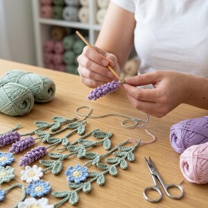 May include: A wooden crochet hook held in hands working on a lavender flower piece. The project features crocheted lavender sprigs, blue and white flowers, and green leaves. Skeins of sage green, lavender, and pink yarn and metal scissors are present.
