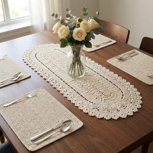May include: An oval cream-colored crochet table runner with a scalloped edge, accompanied by four matching rectangular crochet placemats. A clear glass vase containing a bouquet of pale cream roses and eucalyptus sits in the center.