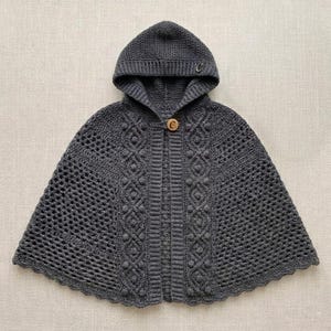 May include: A dark gray hooded cape featuring a textured crochet pattern with a central vertical panel of braided cables and bobble stitches. The garment includes a single wooden button closure at the neckline and a scalloped hem edge.