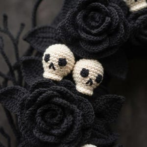 May include: A decorative wreath featuring multiple crocheted black roses with dark green leaves and two small, off-white crocheted skulls with black embroidered eye sockets and nose cavities nestled among the dark floral elements.