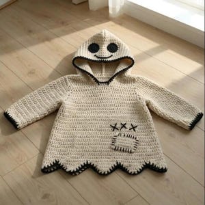 May include: A cream-coloured crochet hooded jumper featuring a ghost-like face on the hood with black circular eyes and a curved mouth. The garment has long sleeves, a black stitched hem, a small square pocket, and three black X-shaped stitches.