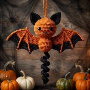May include: A crocheted bat ornament featuring a round orange head and body, black ears, and black-tipped orange wings. The bat has a long, black, spiraled tail and is suspended by an orange yarn loop. It has small black eyes and a stitched smile.