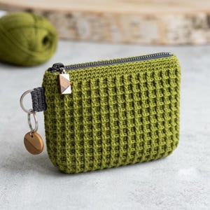 Puede incluir: Un estuche rectangular de ganchillo en color verde oliva con un patrón de punto de gofre texturizado. El estuche tiene una cremallera gris oscuro con un pequeño tirador geométrico de madera y un llavero de metal plateado en una trabilla gris.