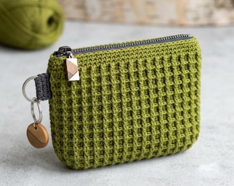 May include: A rectangular olive green crochet pouch featuring a textured waffle stitch pattern. The pouch has a dark gray zipper closure with a small geometric wooden pull, and a silver metal keyring attached to a gray fabric loop with a wooden disc.